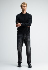 Schwarzes Sweatshirt mit Rundhalsausschnitt und Raglanärmeln, kombiniert mit dunkelgrauen Jeans mit einem ausgewaschenen Muster. Modell trägt schwarze Schuhe.