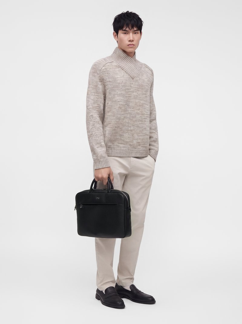 Jeune homme portant un pull beige et un pantalon, tenant une mallette en cuir noir, debout devant un fond clair uni.
