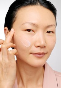 Osoba delikatnie nakłada przezroczysty, lekki krem na policzek, ukazując gładką skórę oraz miękką różową koszulę z kołnierzykiem.