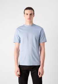 Lyseblå bomulls T-skjorte med korte ermer og høy hals. Stoffet ser glatt ut og har en avslappet passform, brukt sammen med svarte bukser.