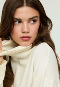 Cremefarbener Pullover mit hohem, umgeschlagenem Rippenkragen. Weicher, strukturierter Strickstoff. Das Model neigt leicht den Kopf und zeigt die Details des Pullovers.