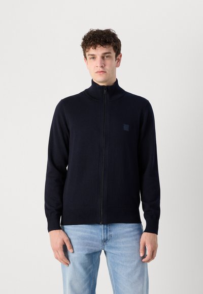 Suéter azul marino con cierre de cremallera, cuello alto, textura de punto y un pequeño parche con logo en el pecho izquierdo. Combinado con jeans azul claro.