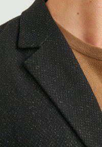 Blazer nero con texture, caratterizzato da un colletto affilato e un sottile motivo intrecciato, abbinato a un maglione marrone a girocollo sotto.