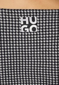 Schwarz-weißer Hahnentrittmuster-Stoff mit einer strukturierten Oberfläche, der ein großes schwarz-weißes "HUGO"-Logo-Etikett trägt.