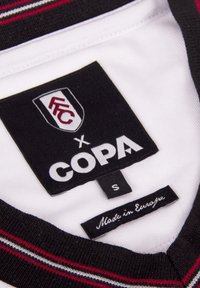 Witte jersey stof met een zwart en rode ribbelkraag. Labels tonen "FFC x COPA," maat "S," en "Gemaakt in Europa."
