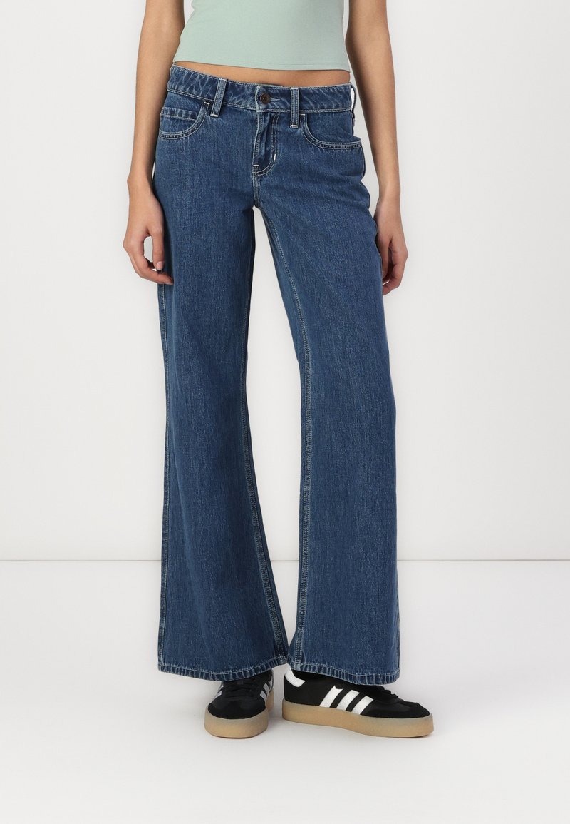 Jeans de ganga azul de perna larga com uma cintura natural, design de cinco bolsos e detalhes de costura subtil. Combinados com sapatilhas pretas.