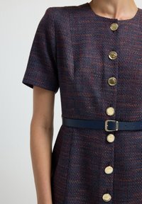 Acercamiento a un vestido de manga corta con textura, grandes botones dorados y un cinturón azul marino oscuro con una hebilla plateada en la cintura.