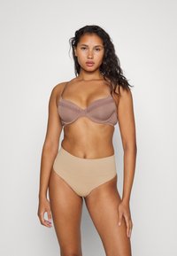 Reggiseno wireless marrone con bordi in pizzo e spalline regolabili, abbinato a mutande beige seamless a vita alta. Texture liscia, toni neutri.