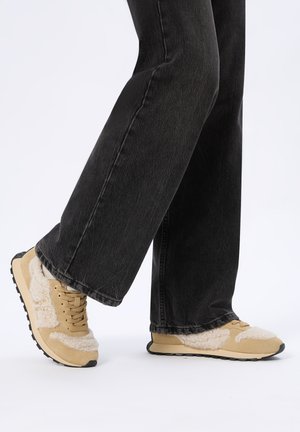 Persona che indossa sneakers in shearling beige e bianco abbinate a jeans neri a gamba larga, in piedi su uno sfondo bianco semplice.