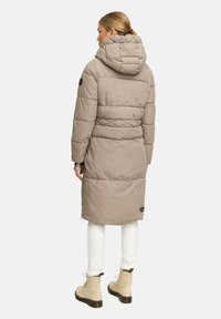 Kvinna iklädd en ljusbrun vadderad lång kappa med huva, vita byxor och beige snörstövlar, vänd bort från kameran mot en neutral bakgrund.