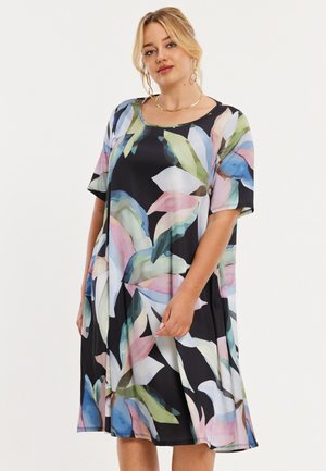 Kvinde iført en sort kjole med store pastel-farvede abstrakte bladmønstre, korte ærmer og rund halsudskæring, stående mod en hvid baggrund.