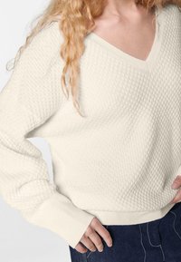 Cremfarbener, strukturierter Strickpullover mit V-Ausschnitt, weiten Ärmeln und gerippten Bündchen. Über dunkler Jeans getragen, zeigt er ein lässiges Design.