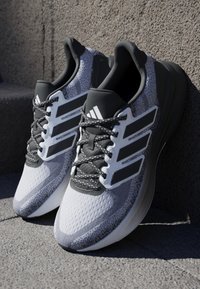 Baskets de sport blanches et grises avec une tige en mesh respirant, une semelle extérieure texturée, des accents noirs et des lacets réfléchissants. Marque à trois bandes visible.