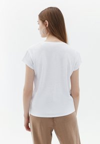 T-shirt bianco a maniche corte con scollatura rotonda. Tessuto morbido, vestibilità rilassata, mostrato di spalle con pantaloni larghi di colore chiaro.