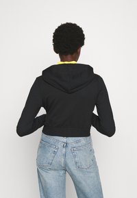 Sudadera corta negra con capucha y cordones, combinada con unos vaqueros de tiro alto de color azul claro. La tela parece suave y con una silueta ajustada.