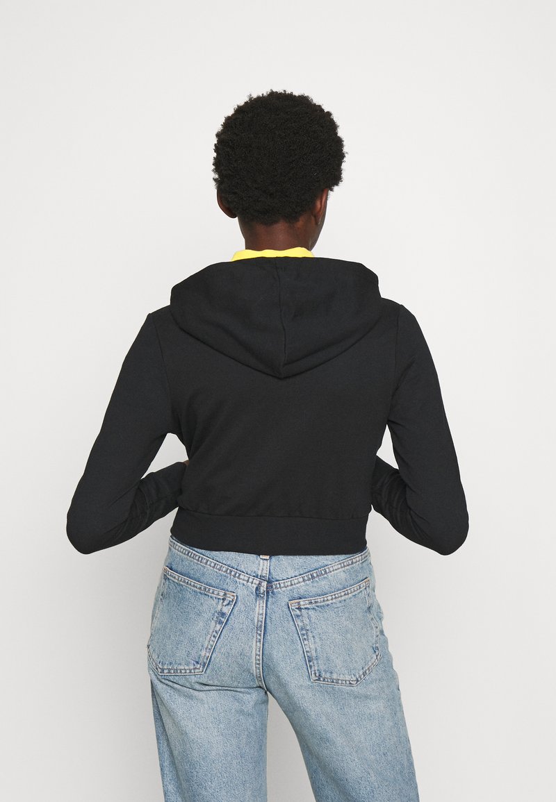Felpa corta nera con cappuccio regolabile, abbinata a jeans chiari a vita alta. Il tessuto sembra liscio con una silhouette aderente.