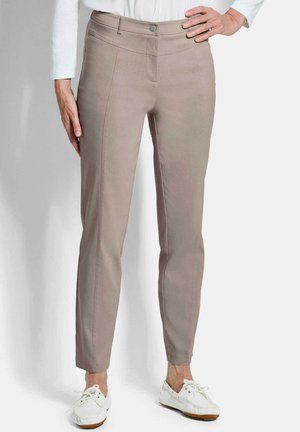 Frau trägt beige eng anliegende Hose und weiße Bootsschuhe, steht mit einer Hand in der Hüfte und der anderen seitlich vor einem einfarbigen Hintergrund.