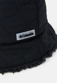 Schwarzer Bucket Hat mit genoppter Textur, ausgestattet mit einem flauschigen Rand und einem rechteckigen Columbia-Logopatch, der an der Seite aufgenäht ist.