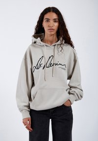 Sudadera con capucha de color beige claro, con bolsillo tipo canguro en la parte frontal y logo negro. Hecha de material suave con un corte relajado. Jeans negros.