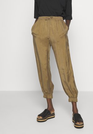Persoon draagt losse beige broek met trekkoord en elastische boorden en zwarte platformsandalen, staand tegen een effen lichte achtergrond.