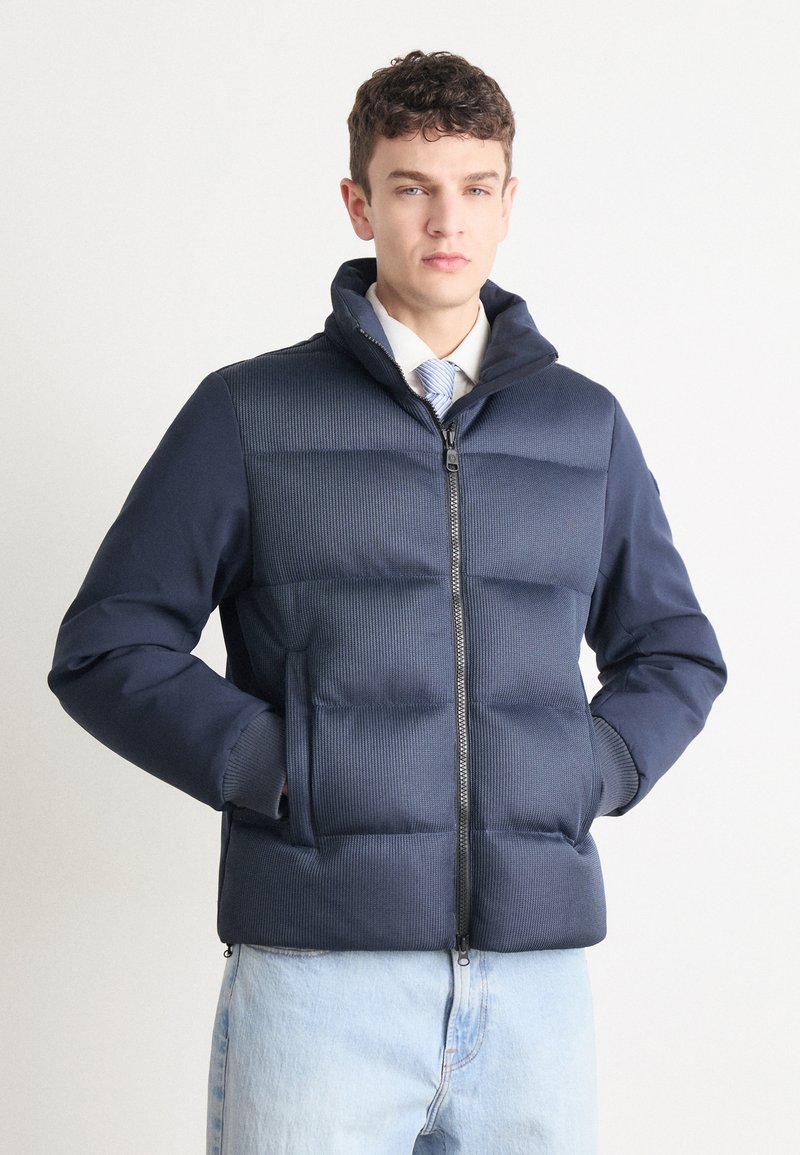 Μπλέ πούφο μπουφάν με φερμουάρ μπροστά, υφαντό επάνω πάνελ, ribbed μανίκια και διπλές πλευρικές τσέπες, συνδυασμένο με ανοιχτό χρώμα τζιν παντελόνι.