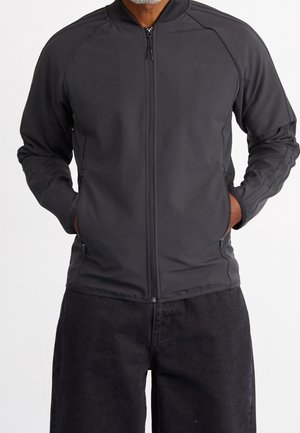 Homme portant une veste noire zippée intégrale, mains dans les poches et pantalon noir, debout devant un fond blanc uni.