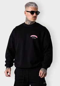 Sweat-shirt oversized noir en tissu doux, avec des textes graphiques et des détails de logo roses. Inclus : poignets côtelés et col rond.