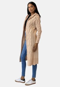 Beige stickad kofta med luva, med flätmönster och ribbade muddar och nederkant. Kombinerad med blå smala jeans och sneakers.