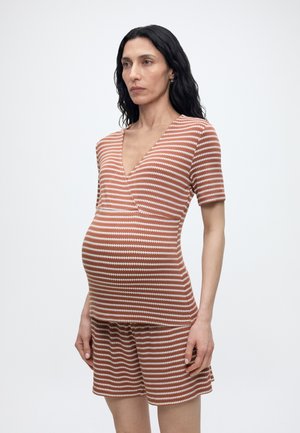 Gravid kvinde med langt sort hår iført en brunt og hvidt stribet kortærmet top og matchende shorts, stående mod en ensfarvet baggrund.