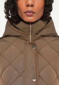 Casaco marrón acolchoado com capuz, apresentando uma gola alta, fecho de correr dourado e botões de pressão. Textura suave com contrastes.