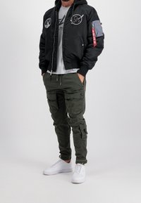 Musta pommitakki, jossa on kangasmerkkejä, harmaa t-paita, oliivinvihreät cargo-housut ja valkoiset lenkkarit. Takissa on vetoketjullinen suljin ja resorireunukset hihansuissa.