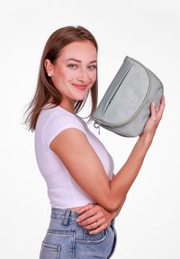 Světle šedá kosmetická taška s zaoblenými okraji, horním zipem a decentním logem na přední straně, kterou drží osoba na bílém pozadí.