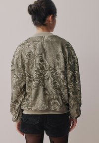 Beaded bomber jacket in a metallic silver hue; features intricate floral patterns and a ribbed collar; paired with dark shorts.