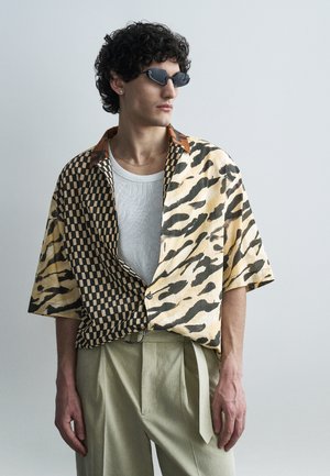 Muž kučeravých vlasov so čiernymi slnečnými okuliarmi, v polovične károvanom a polovične tigrom vzorovanom tričku cez biele tričko a béžových nohaviciach na jednoduchom pozadí.