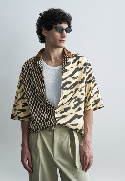 Man with curly hair wearing black sunglasses, a half checkered half tiger print shirt over a white top, and beige pants against a plain background.