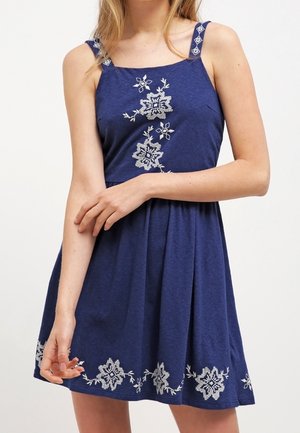 Blauwe mouwloze jurk met witte bloemrijke borduursels op de borst, bandjes en zoom, gedragen door een persoon met gekruiste armen aan de voorkant.