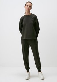 Dunkelgrüner Sweatshirt mit rundem Halsausschnitt und langen Ärmeln, kombiniert mit passenden tapered Jogginghosen und weißen Sneakers. Glatte Textur.