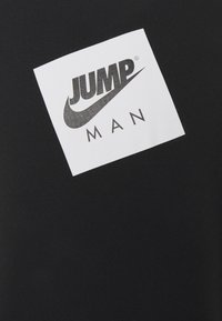Černé tričko s bílým obdélníkovým logem, na kterém je nahoře napsáno "JUMP" a dole "MAN" černým písmem, s důrazem na design swoosh od Nike.