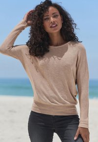 Beige Strickpullover mit langen Ärmeln, lockerem Schnitt, glatter Textur und Bootsausschnitt, kombiniert mit dunklen Jeans. Der Hintergrund zeigt einen Strand.