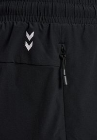 Close-up van zwarte stoffen broek met een elastische tailleband, een verticale ritszak en twee witte chevron-logo's boven de zak.