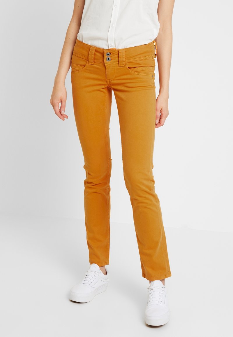 Personne portant un pantalon slim couleur moutarde, des baskets blanches et une chemise blanche sans manches boutonnée, debout devant un fond uni.