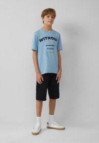 Hellblaues Baumwoll-T-Shirt mit fettem schwarzen Text "OHNE STANDARDS" und Grafik darunter, kombiniert mit schwarzen knielangen Shorts und weißen Turnschuhen.