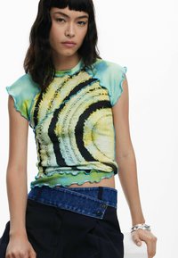 Bunt gestreifter Strickoberteil mit einem wirbelnden Muster in Grün, Gelb und Schwarz und gewellten Kanten. Kombiniert mit dunklen Hosen und Denim-Accessoires.