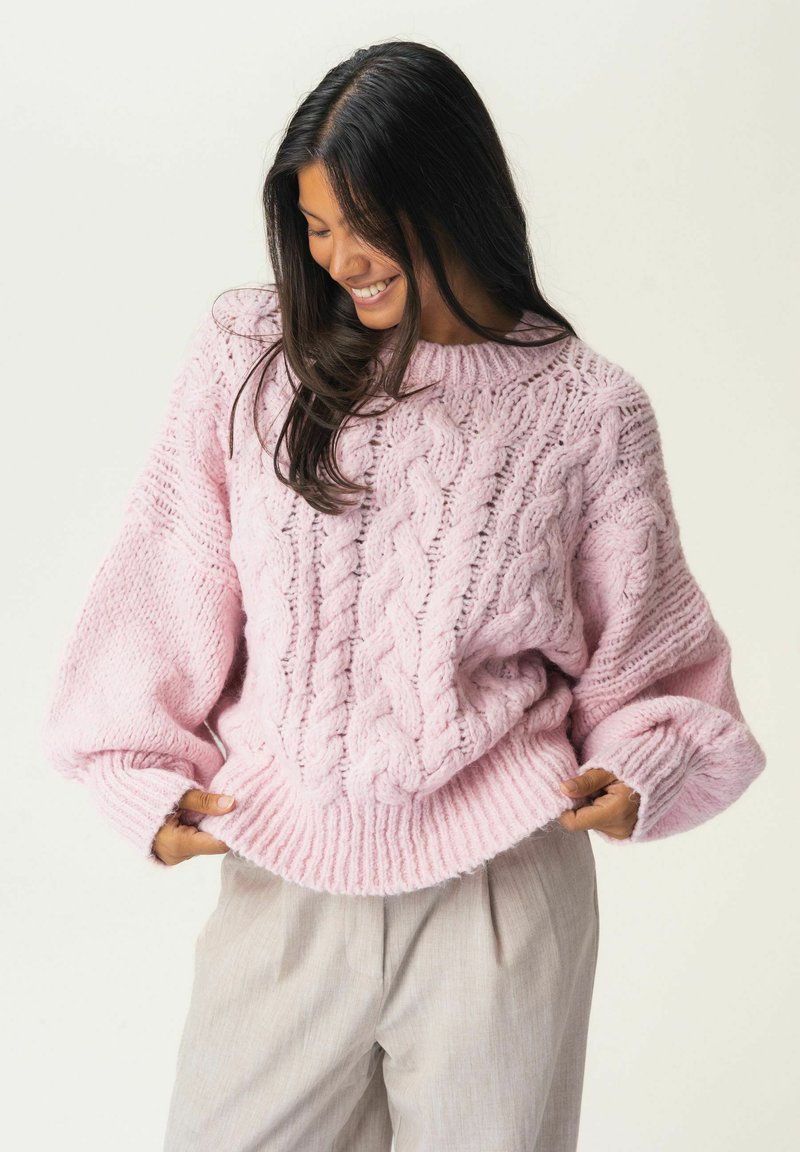Pull en tricot rose clair avec un motif en cable, manches larges et ourlet côtelé. Le tissu semble doux et épais, favorisant la chaleur.