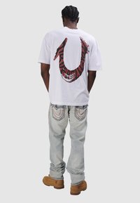 Biała, oversize'owa koszulka z dużą czerwoną i czarną grafiką na plecach, zestawiona z jasnoniebieskimi dżinsami z kontrastującym ściegem i brązowymi botkami.