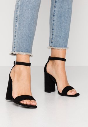Füße, die schwarze Wildleder-Sandalen mit Blockabsatz und Knöchelriemen tragen, kombiniert mit hellblauen Jeans mit ausgefranstem Saum, auf einem weißen Boden.