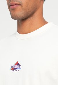 Λευκό βαμβακερό t-shirt με στρογγυλή λαιμόκοψη. Διαθέτει πολύχρωμο λογότυπο "tommy" πάνω από το "JEANS" σε μωβ χρώμα. Απαλή υφή και άνετη εφαρμογή.