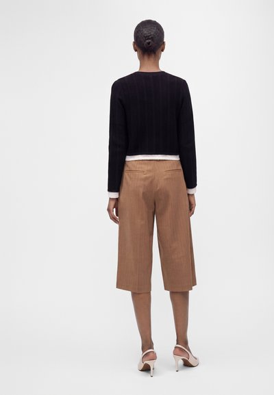 Femme aux cheveux noirs en chignon portant un haut noir à manches longues, des culottes beiges et des chaussures blanches à talons hauts, debout de dos sur un fond uni.