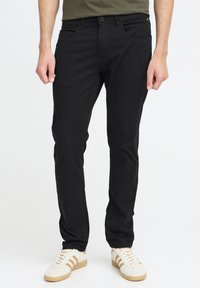 Pantalon en denim noir à coupe slim, avec cinq poches et une fermeture à boutons. Porté avec des baskets blanches à rayures beiges.