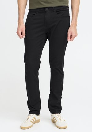 Schwarze Denim-Hose im Slim-Fit mit fünf Taschen und einem Knopfverschluss. Kombiniert mit weißen Sneakers mit beigen Streifen.